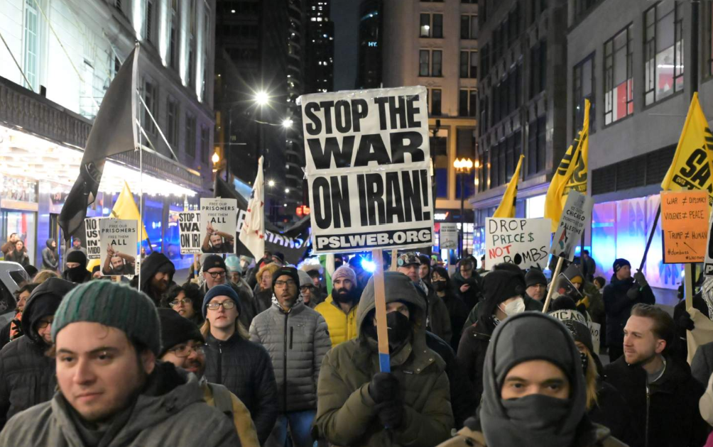 A protest crowd in an urban setting at night. A large sign that says "Stop The War On Iran! PSLWEB.ORG" is centered. Protesters are wearing winter garments.