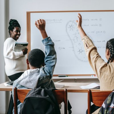 Two students raise their hands in a classroom. Photo courtesy Katerina Holmes on Pexels.