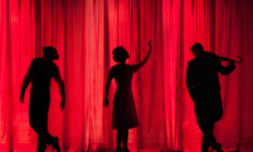 Three silhouettes stand in front of a red curtain