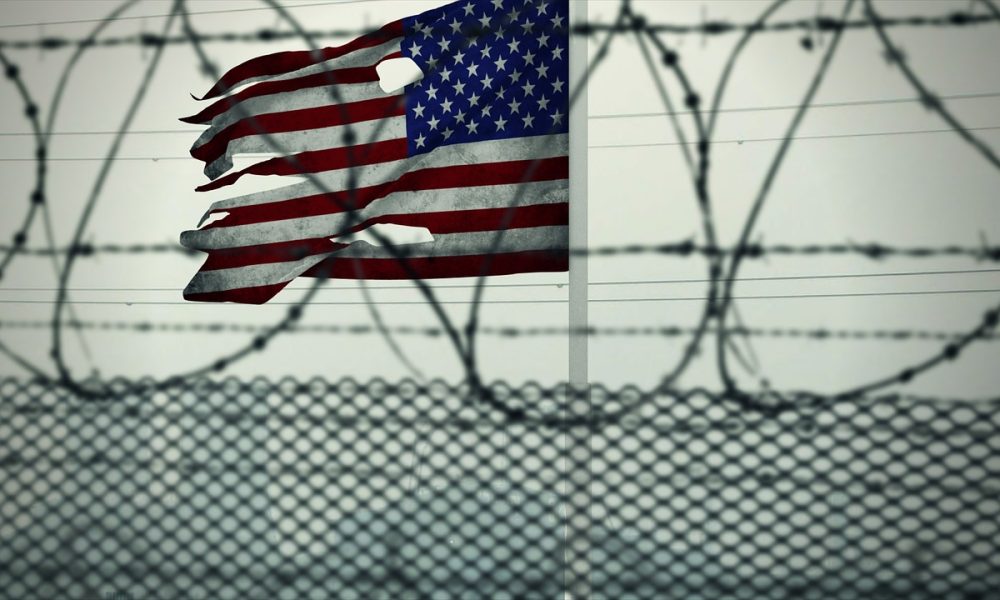American Flag with holes in it behind a barbed wire fence. Photo courtesy of TayebMEZAHDIA on Pixabay.