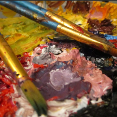 A close up image of a painter's palette covered in dried and wet paint of many colors, with three paint brushes laid on top.