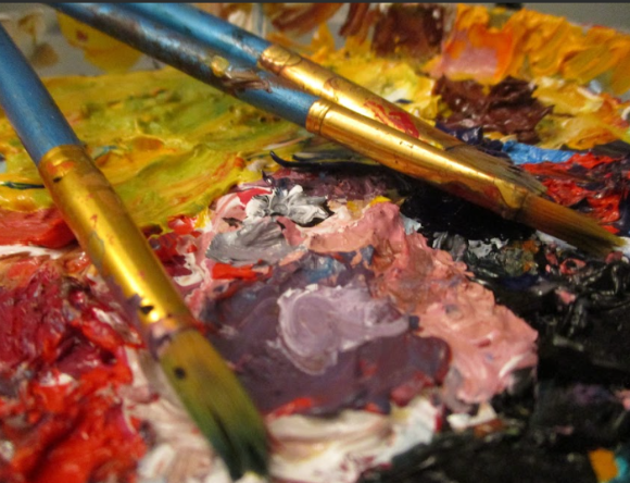 A close up image of a painter's palette covered in dried and wet paint of many colors, with three paint brushes laid on top.