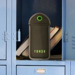 A photo of the Yondr phone pouch, a grey plush case with bright green accents, situated in a blue school locker propped up by books