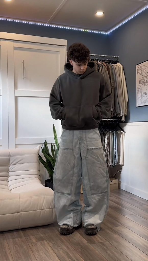 Instagram fashion post of a teen boy wearing a cropped brown hoodie, grey baggie jeans, and black shoes.