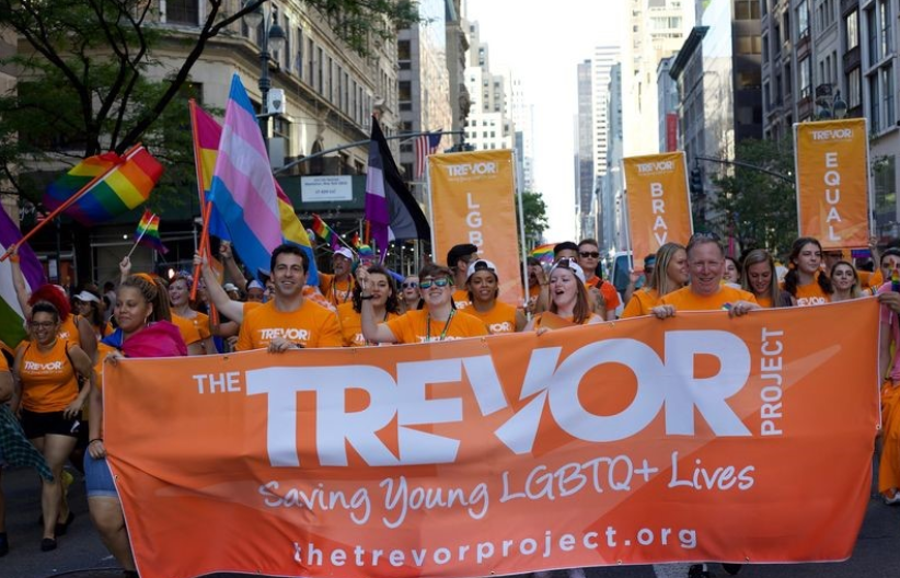 People at a protest in a metropolitan area, carrying an orange banner with the Trevor Project's name and logo.