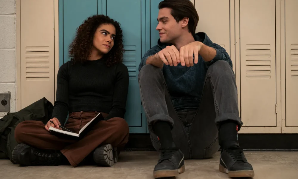 A teenage couple sitting on the floor of a high school with their backs to the lockers.