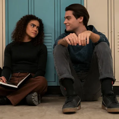 A teenage couple sitting on the floor of a high school with their backs to the lockers.