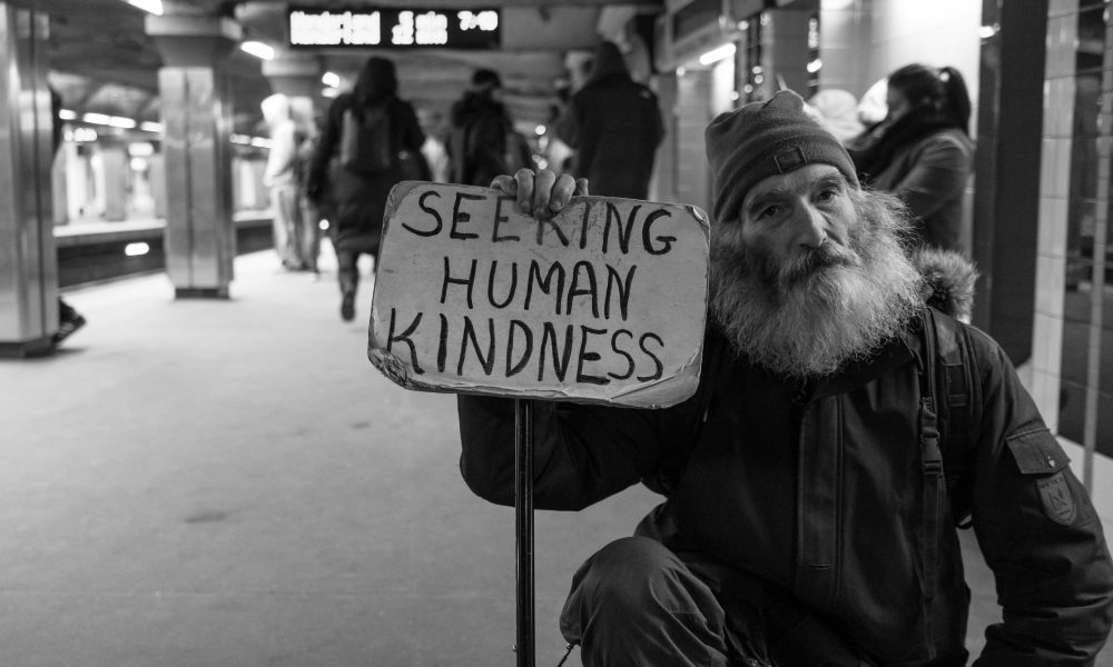 Black and white image of an unhoused person with a sign that reads Seeking Human Kindness.