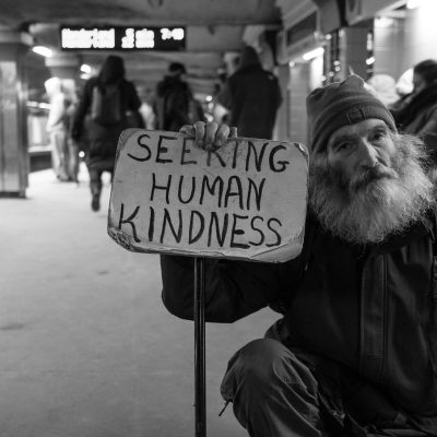 Black and white image of an unhoused person with a sign that reads Seeking Human Kindness.