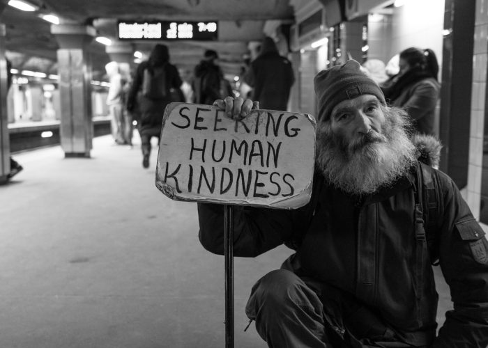 Black and white image of an unhoused person with a sign that reads Seeking Human Kindness.