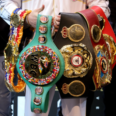 Multiple large boxing champion belts draped on the arms of a man dressed in white.