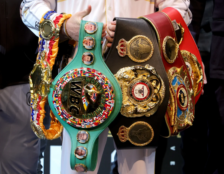 Multiple large boxing champion belts draped on the arms of a man dressed in white.