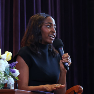 Image of Ayo Edebiri speaking into a microphone at a panel discussion / interview. Seated next to flowers.