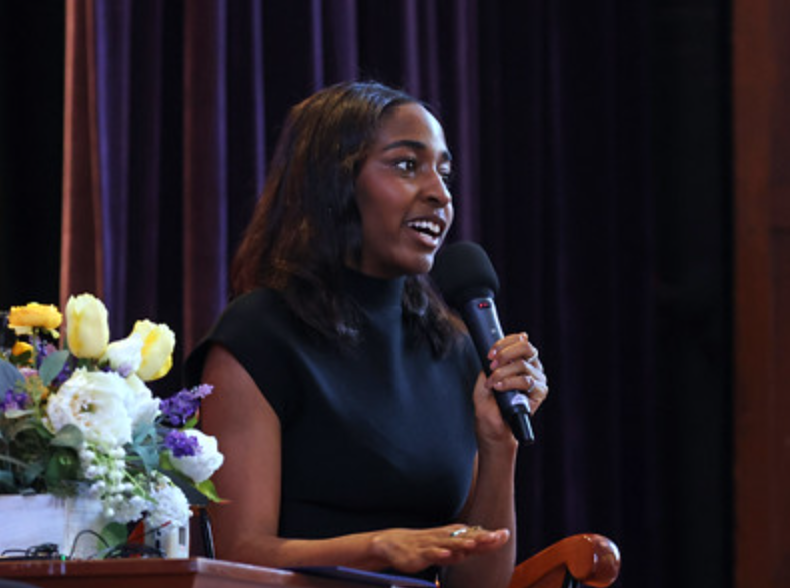 Image of Ayo Edebiri speaking into a microphone at a panel discussion / interview. Seated next to flowers.
