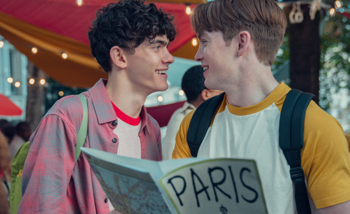 Happy image of a queer couple from the show Young Royals looking at a book that says Paris on it.