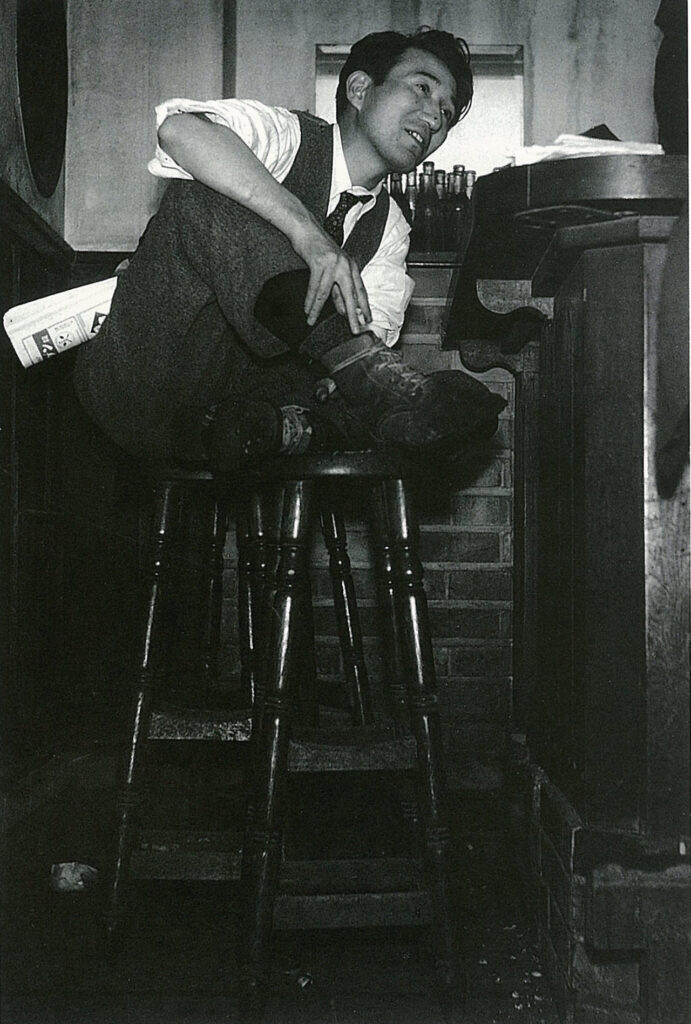 Author Osamu Dazai perched on a bar stool with his feet on another bar stool. Wearing 1930s style vest, tie, white shirt, and slacks.