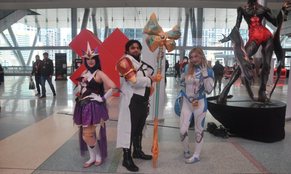 3 cosplayers posing in a convention hall lobby. From left to right is a woman dressed as Syndrome's Star Guardian Skin from League of Legends, a man dressed as Jayce from Arcane, and a woman dressed as Sue Storm from Fantastic Four.