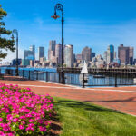 A brick walkway in East Boston's Pier's Park invites you to stroll past the lush green grass and bright flowers to the black wrought-iron railing by the waterfront. Gaze out across the Boston Harbor to see sailboats gliding by or the numerous buildings of all sizes in downtown Boston. With the clear blue sky, lights are not needed, but decorative street lamps are ready for nightfall.